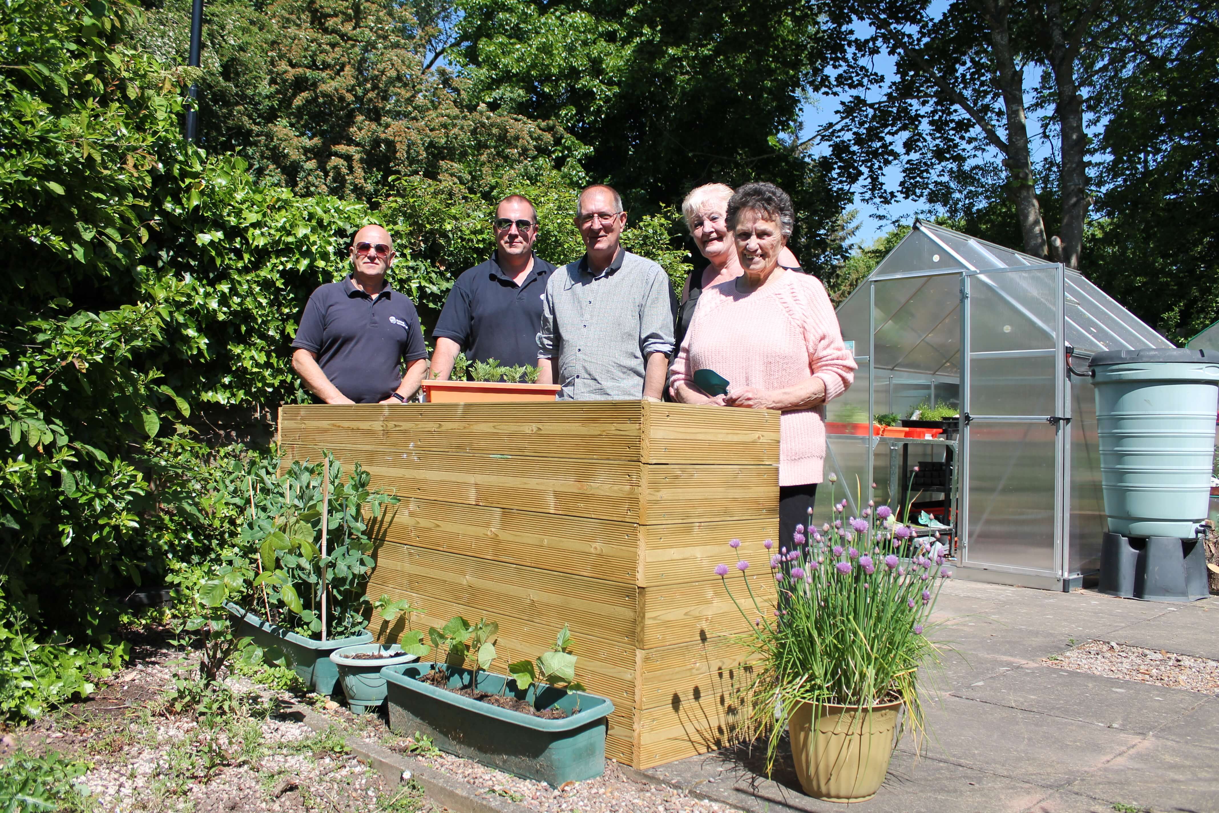 three men and two women standing in a garden in front of a planter box in the sunshine smiling holding gardening tools with a small glasshouse behind them