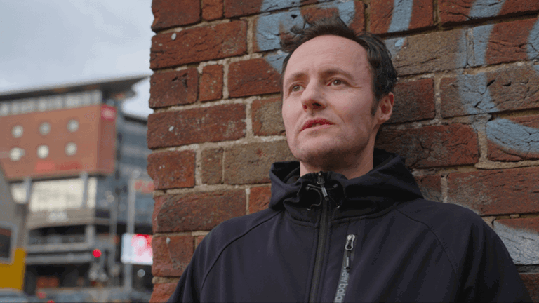 upper body view of a man named Kieran wearing a zipped up black jacket leaning against a brick wall staring into the distance